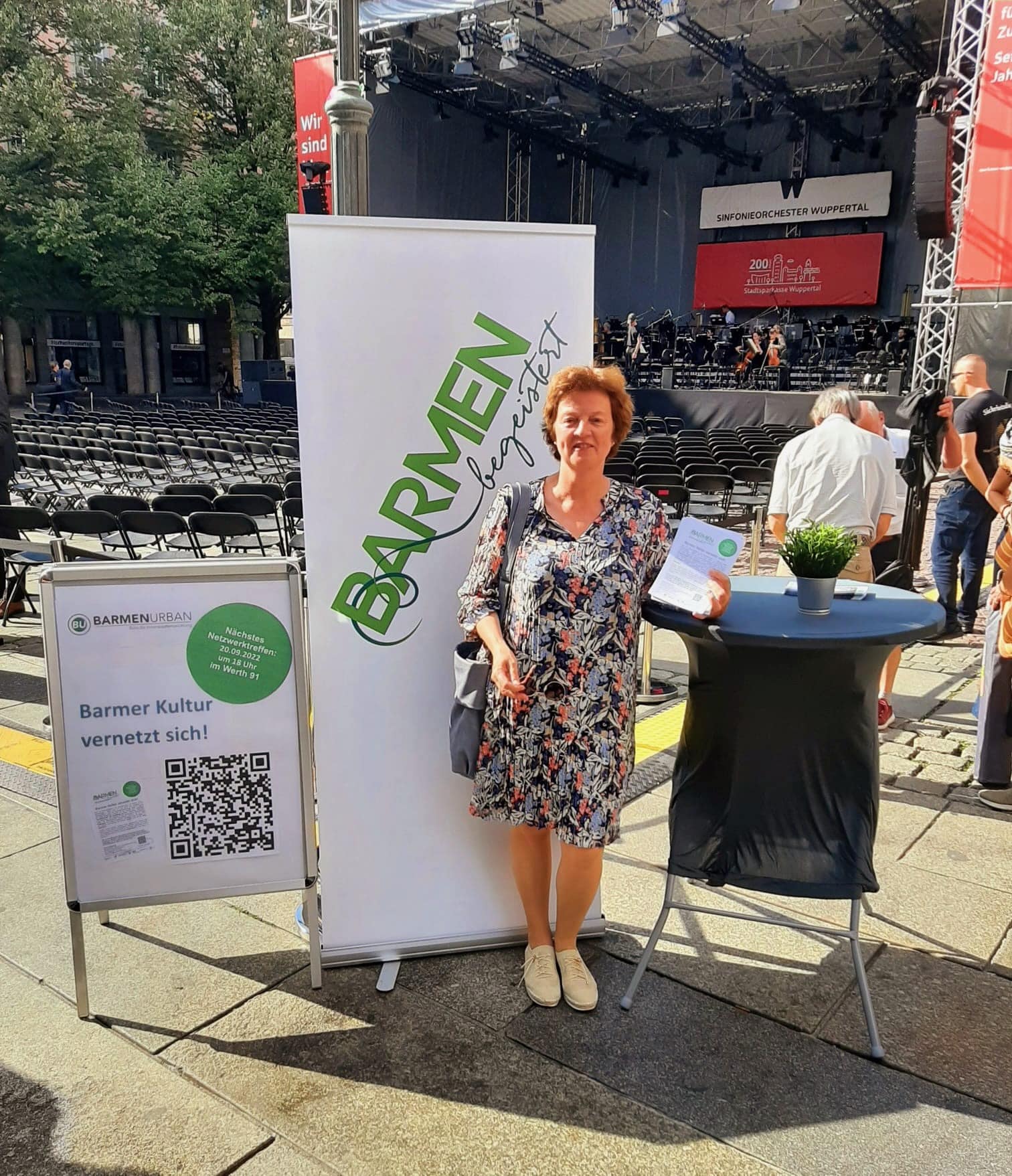 2022 Dörte Baldsteht neben Aufsteller mit Netzwerkinformationen, 'Barmen besteister' Plakat und Stehtisch, im Hintergrund eine Bühne mit Stühlen auf dem Johannes-Rau-Platz