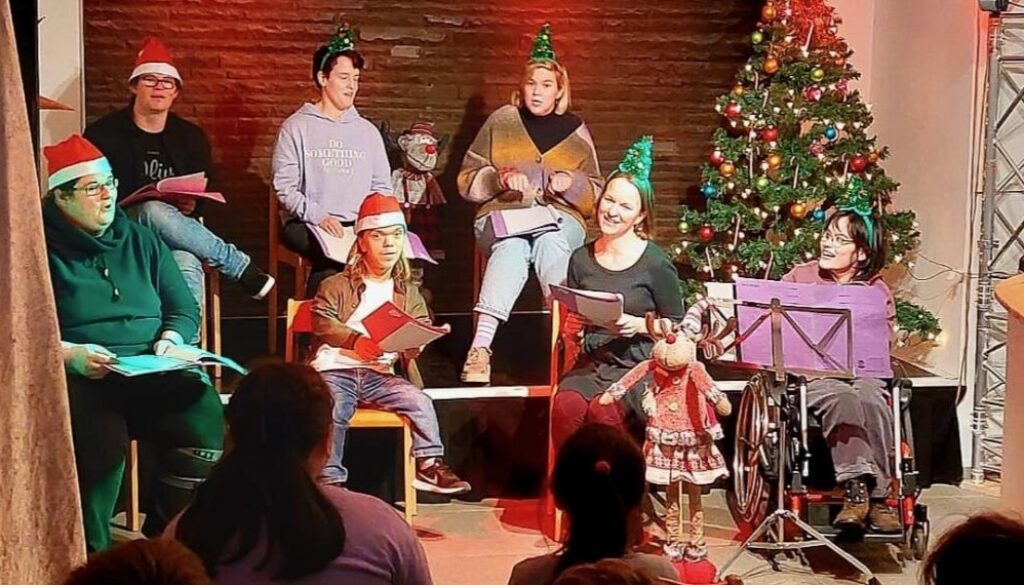 Eine Gruppe von Menschen sitzt mit Weihnachtsmützen neben einem geschmückten Tannenbaum, im Publikum sitzen Kinder