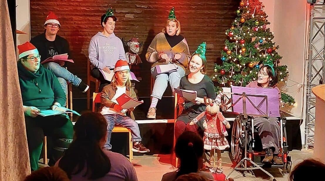 Eine Gruppe von Menschen sitzt mit Weihnachtsmützen neben einem geschmückten Tannenbaum, im Publikum sitzen Kinder