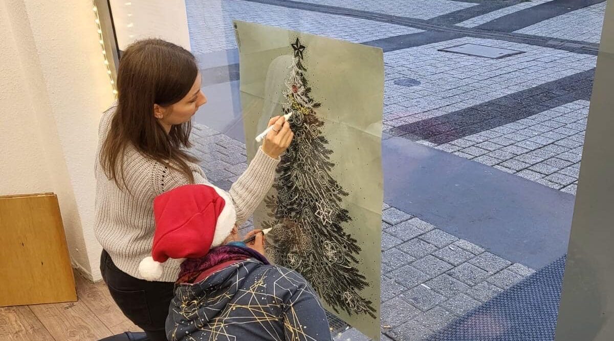 Zwei Frauen, eine davon mit Weihnachtsmannmütze, hocken vor einem Fenster und malen einen Tannenbaum auf die Scheibe