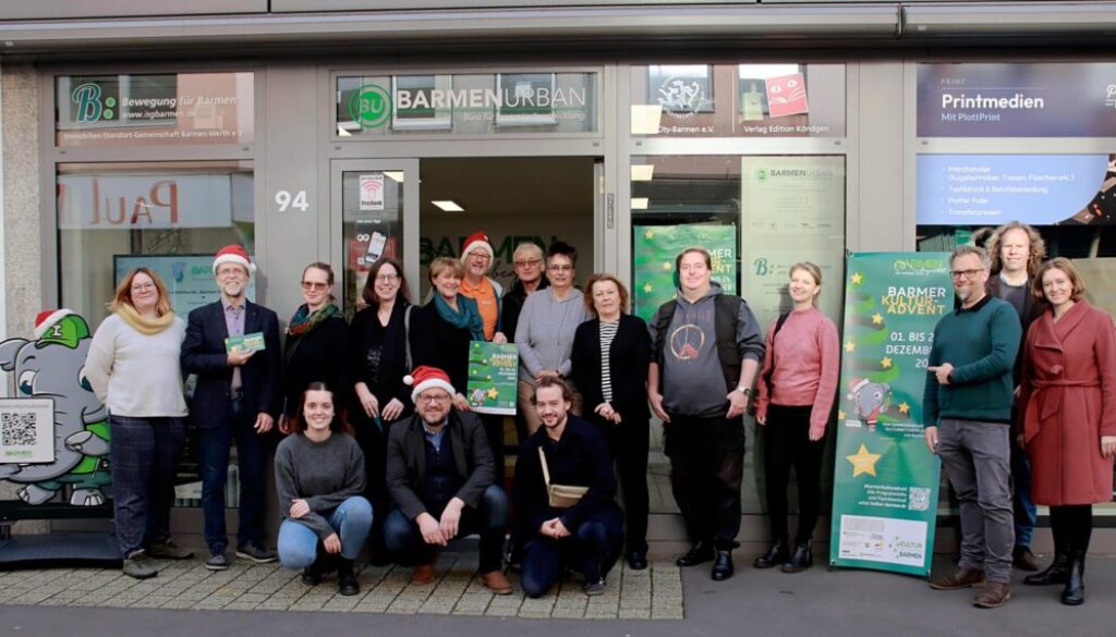 Gruppe von Menschen posiert für ein Foto vor dem BarmenUrban Büro, einige tragen Nikolausmützen, neben ihnen ein grünes Banner zum Kulturadvent Barmen 2024 und ein Aufsteller mit einem Elefanten