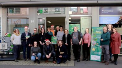 Gruppe von Menschen posiert für ein Foto vor dem BarmenUrban Büro, einige tragen Nikolausmützen, neben ihnen ein grünes Banner zum Kulturadvent Barmen 2024 und ein Aufsteller mit einem Elefanten