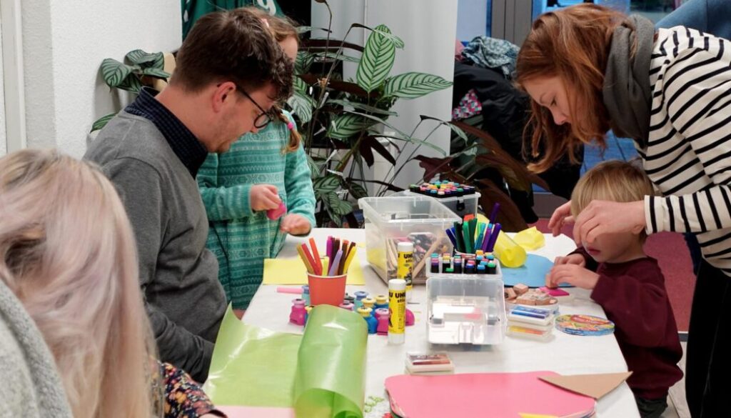 Erwachsene und Kinder sitzen am Tisch der mit Bastelmaterial bedeckt ist